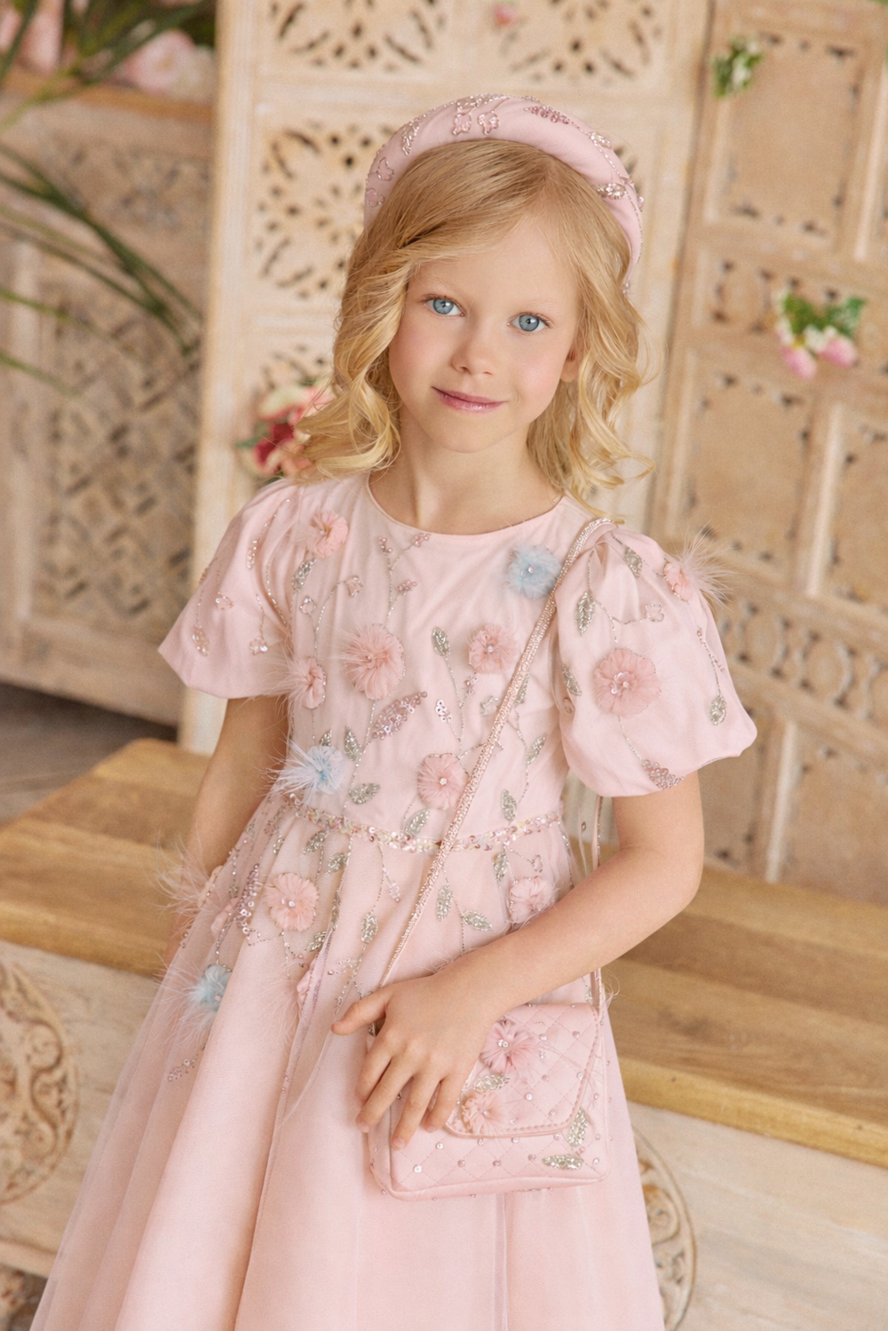 Young girl in a pink floral dress standing in front of a decorative wooden wall.