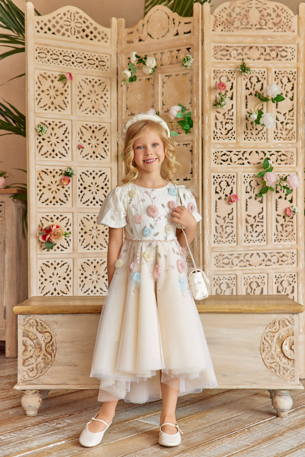 Young girl smiling in a floral dress with a decorative headband and bag, sitting on a wooden bench.