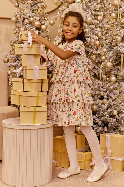 Young girl in a floral dress standing next to a stack of gift boxes with a decorated Christmas tree in the background.