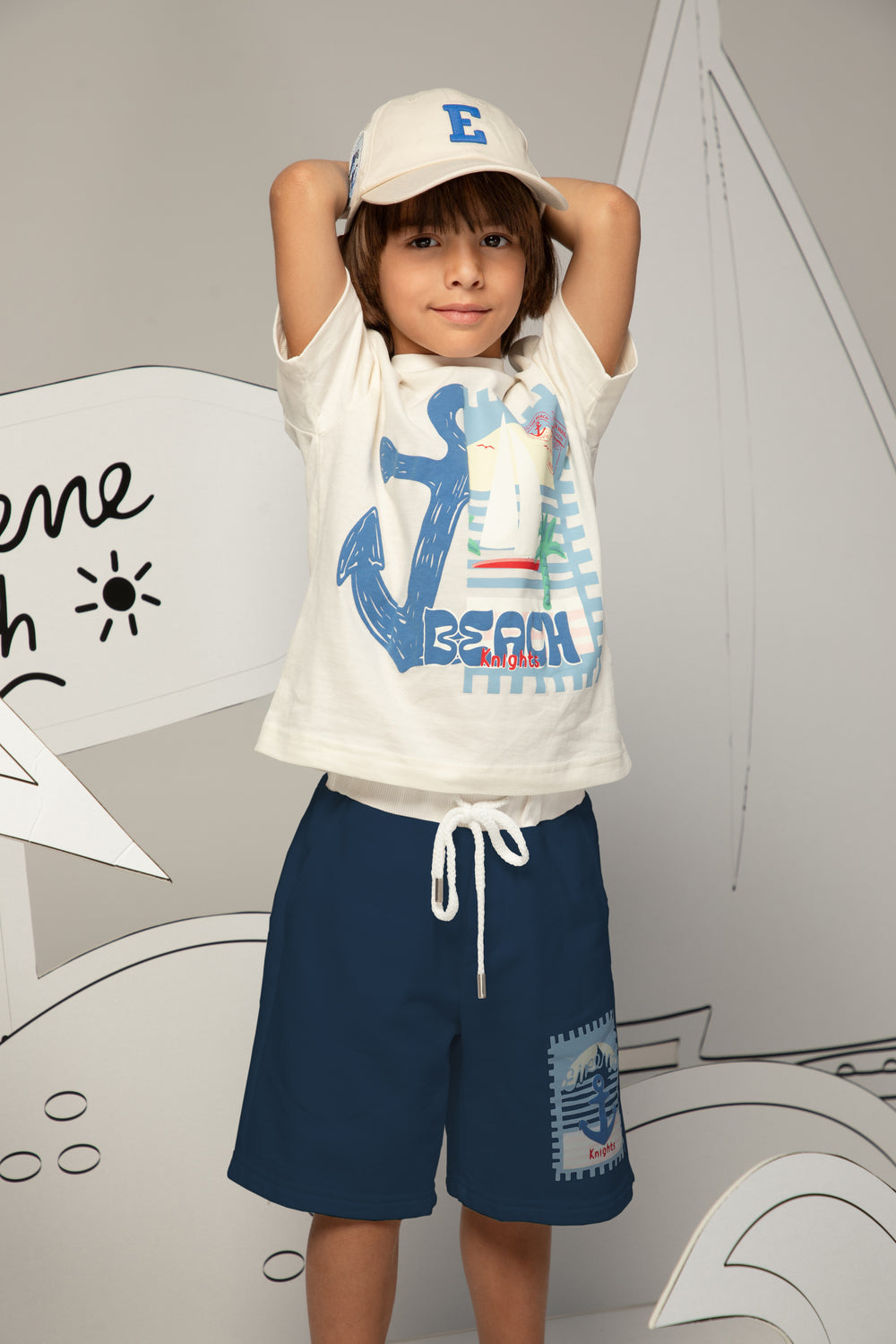 Child wearing a white t-shirt with a blue anchor design and navy shorts, standing against a gray background.